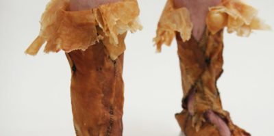 A photograph of a footwear art piece on a pair of legs. The footwear is made out of a brown leather material, and thread together with black thread. On the top of the leather footwear, the leather is folded over in a lighter brown color.