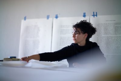 A young woman in the process of printmaking. She has an outstretched hand and is looking towards the prints on the table in front of her.