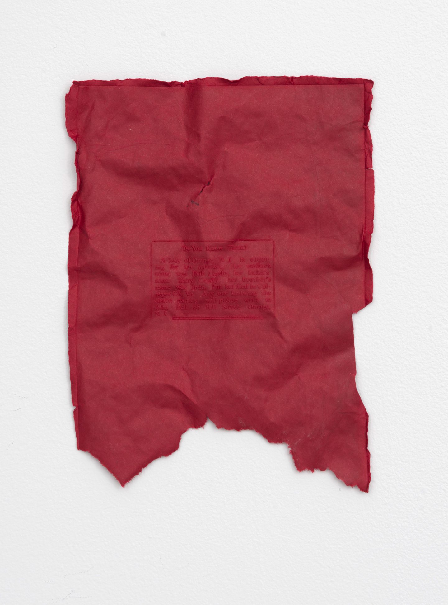 On a white wall, a single piece of crumpled and torn red paper.